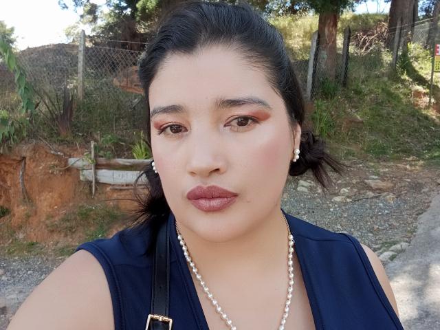 ZafiRouge has dark hair, brown eyes, and full lips with pink lipstick. She's wearing a navy blue top, pearl earrings, and a pearl necklace. Her ass is not visible.