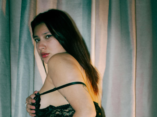 LadyStarOne con lunghi capelli scuri, indossa lingerie di pizzo nero, tiene le mani sui seni, guarda seducentemente la fotocamera, con una tenda sullo sfondo, illuminazione verde e gialla tenue..