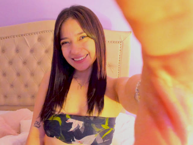 CattsyCathy with long black hair, smiling, wearing a strapless floral top, showing a tattoo on her right arm, taking a selfie, her hand in the foreground.
