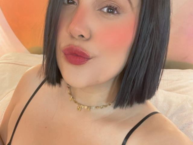 SamantaMuller with black bob, pink blush, red lipstick, black strap, gold necklace, and cute pout. Her tits are small, and she's looking at the camera.
