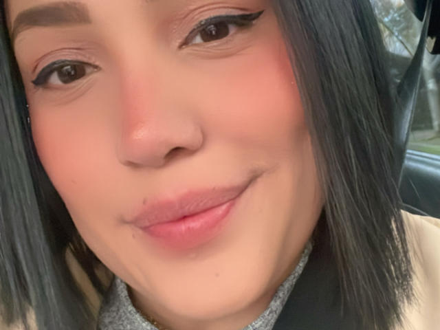 SamantaMuller has a cute face with black eyeliner, pink blush, and black hair, smiling with pink lips and a small nose, wearing a grey and black top.