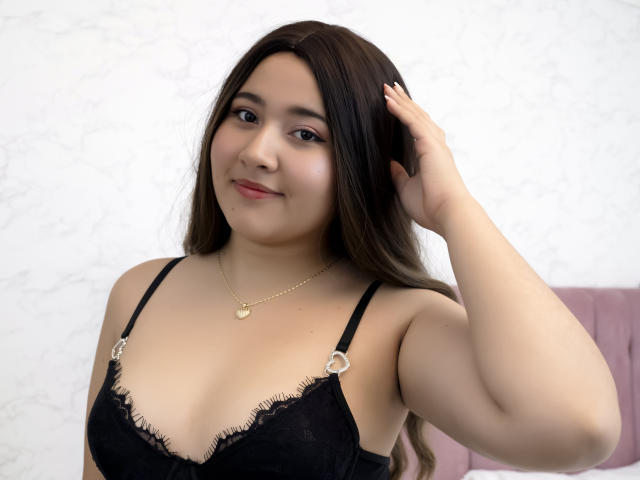 CatalinaRosel with long brown hair, black lace bra with fringed edges, gold necklace, hand in hair, smiling, fair skin, medium tits, soft light, pink headrest in background.