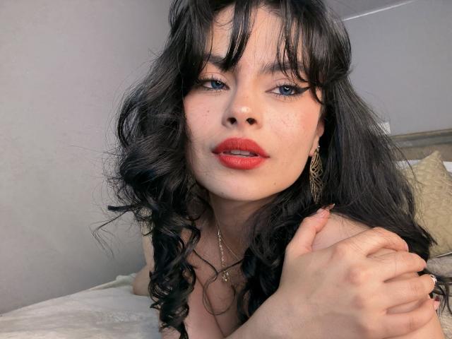 ClariceMontclair with wavy black hair, red lipstick, and blue eyes, holding her chest, showing a hint of her tits, wearing gold earrings, in a soft light, looking at camera.