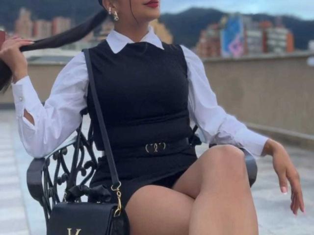 Laila Barker met een zwarte jurk, wit shirt, zwarte riem, gekruiste benen, haar in staarten, zwarte tas, zittend op een versierde stoel, haar achterwerk zichtbaar, rode lipstick, stadsachtergrond..