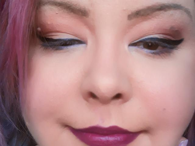 MissCristina's face with pink hair, dark eyeliner, brown eyes, and purple lipstick. Her nose is centered, and she's giving a subtle, confident smile. Her skin is fair.
