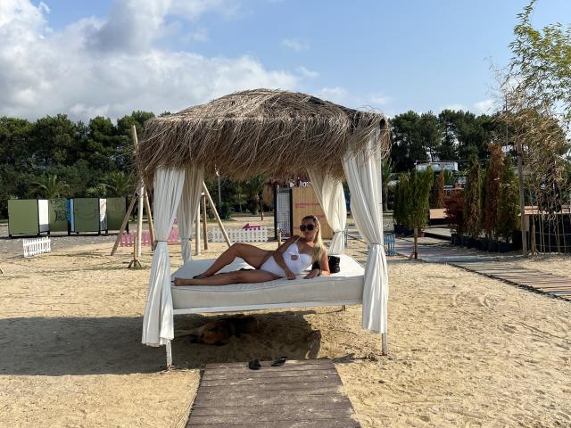 AmandaSexyX è sdraiata su un letto bianco sotto una tettoia di paglia, indossa un bikini bianco, con i seni e i glutei visibili, e tiene in mano una tazza nera..
