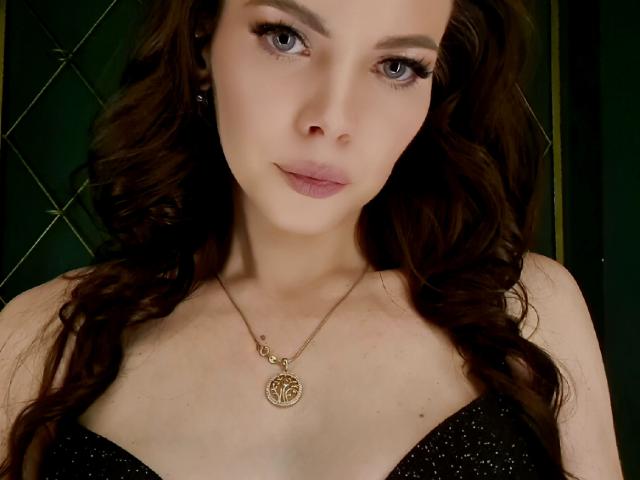 TinaPeachX with long brown wavy hair, blue eyes, and pink lipstick, wearing a black sparkly top and gold necklace with a circular pendant, looking straight at the camera.