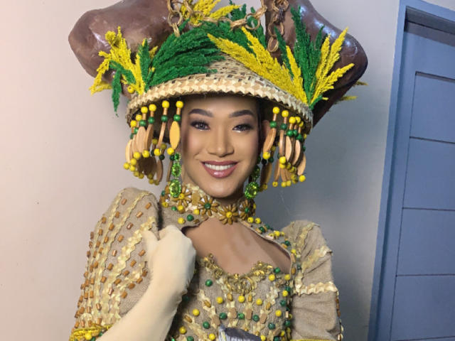 AsianHugeCockxX in a colorful, ornate headdress with yellow and green feathers, wearing a bejeweled, gold and green dress, smiling with visible makeup and white glove.