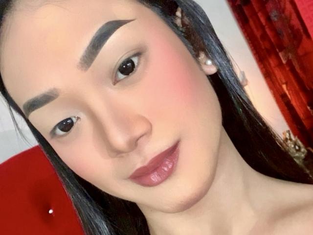 AsianHugeCockxX with thick black eyebrows, pink blush, glossy pink lips, and long black hair, wearing pearl earrings, smiling slightly, with a red background.