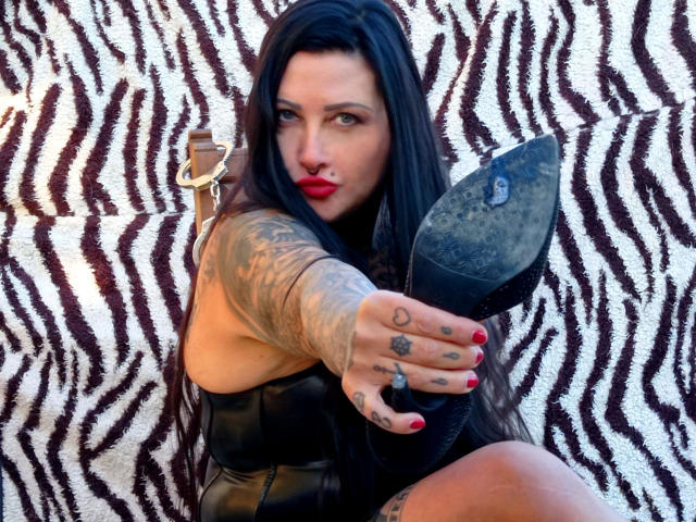 StrengeTante-hot with long black hair, tattoos, red nails, and black leather, points at a dirty shoe with a pensive expression, full red lips, and a nose ring.