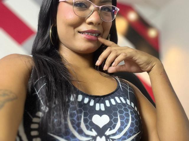 ChloeBrownOne with glasses, dark hair, and hoop earrings, biting her finger, wearing a hexagon-patterned black top, showing her tits, smiling softly, with a tattoo on her arm.