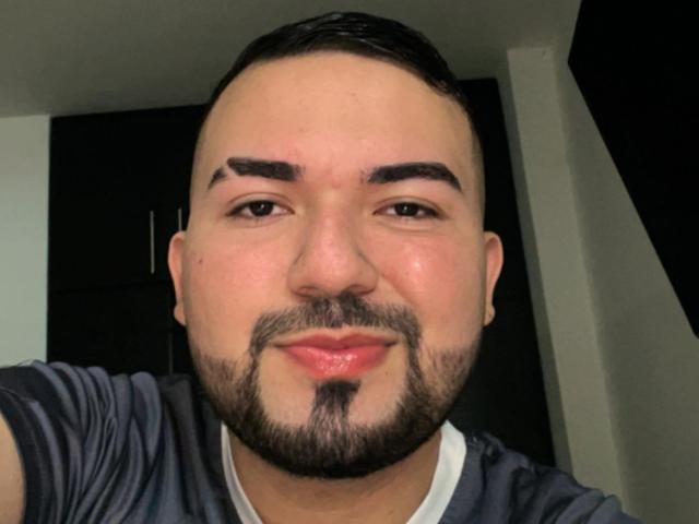 GaelB a un visage rond, des sourcils foncés, des cheveux noirs courts et une barbe avec une moustache. Il porte une chemise grise avec un col blanc..