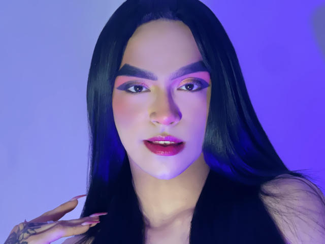 MelissaHotX69 has long black hair, pink and purple eyeshadow, thick eyebrows, glossy pink lips, and a tattoo on her right hand, with blue and purple lighting.