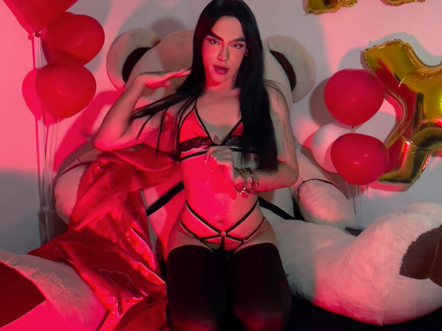 MelissaHotX69 with long black hair, wearing black lace lingerie and thigh-high stockings, sitting on a giant stuffed animal, surrounded by red balloons and gold number two balloons.