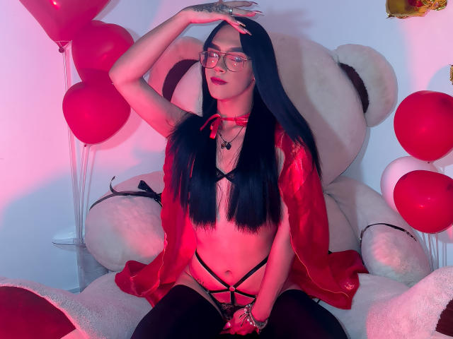 MelissaHotX69 with long black hair, glasses, red choker, red cape, black strappy lingerie, and black stockings, sitting on a giant white teddy bear with red balloons.