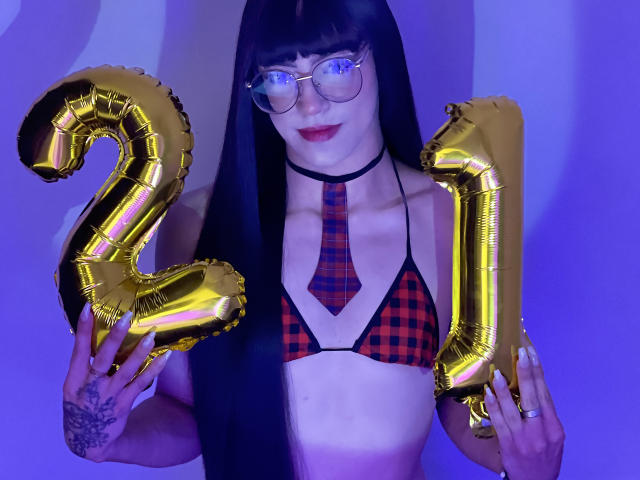 MelissaHotX69 with long black hair, glasses, red plaid bikini, black choker, gold "2" and "1" balloons, showing tattoos, holding balloons, purple lighting, smiling, wearing red plaid tie.