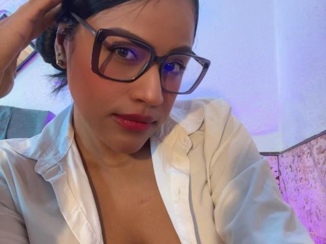 IvannaDuran with glasses, red lips, and white shirt, showing cleavage, right hand in hair, medium skin, black hair in a bun, subtle smile, purple-blue light.