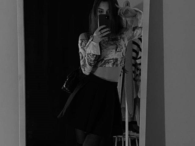 JeniferStars in a black and white mirror selfie, wearing a long-sleeve, patterned crop top and a high-waisted, dark skirt, holding a phone, with a bag on her shoulder.
