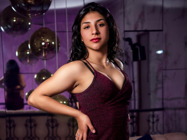 TatianaMorelli with medium tits, wearing a sparkly red dress, has dark wavy hair, red lipstick, and a confident look, standing with one hand on her hip.