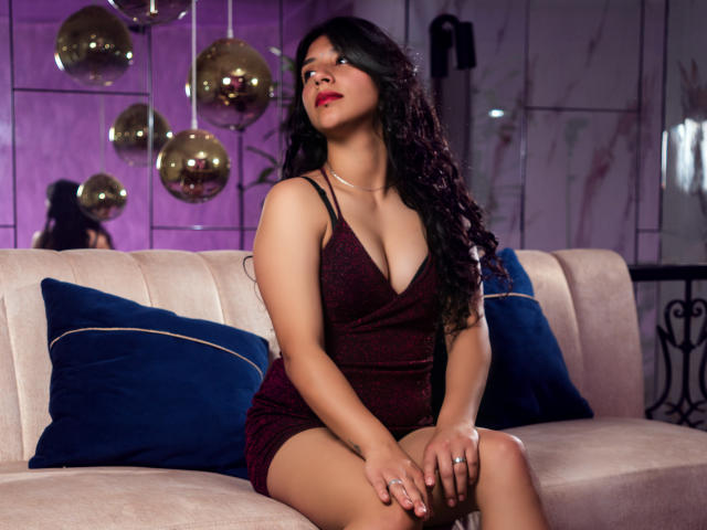 TatianaMorelli sits on a beige couch, wearing a sparkly red dress, showing her boobs, with long curly black hair, red lipstick, and blue pillows on both sides.