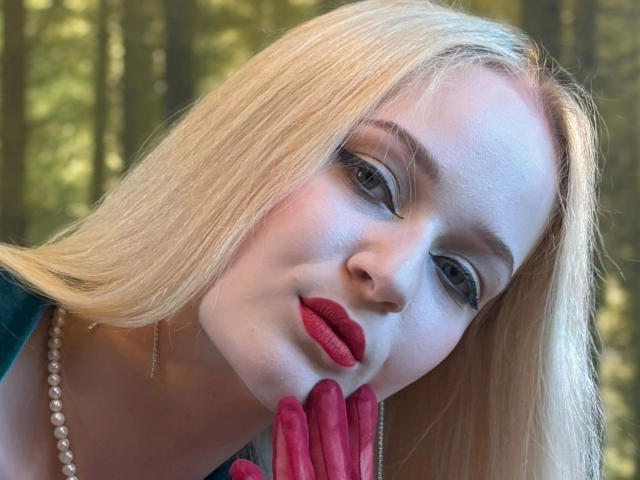 SophiaLust - belle, avec des cheveux blonds, du rouge à lèvres rouge et des gants rouges, légèrement penchée, la main sur le menton, portant un collier de perles, dans une lumière forestière douce..