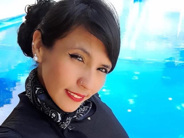 DaniaHot69 with black hair, red lipstick, black scarf, and black shirt, smiling at the camera, wearing a nose ring and earrings, with blue background.