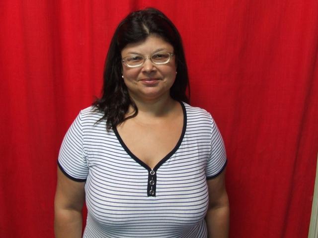 A SweetKarinaX tem seios de tamanho médio, vestindo uma camisa listrada branca e preta com um zíper preto, óculos e cabelo ondulado escuro, sorrindo em frente a uma cortina vermelha..