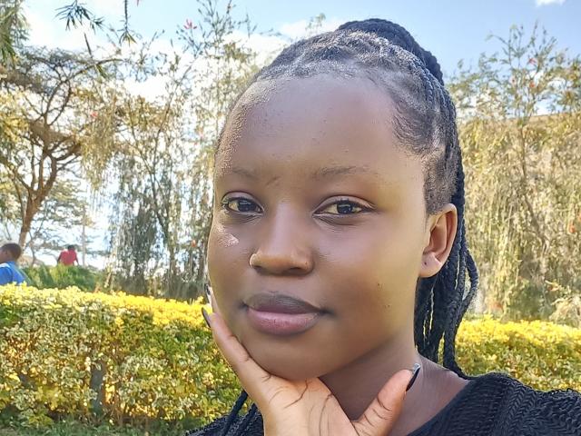NellyVane's face, dark brown skin, small nose, full lips, black braided hair, slight smile, hand under chin, wearing black top, outdoors, blue sky, green bushes, sunlit.