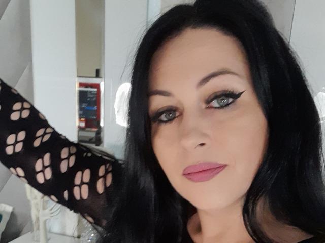 Sellhena, com cabelo preto, olhos verdes e batom rosa, vestindo uma roupa preta com mangas recortadas, olhando para a câmera com um leve sorriso. Sua maquiagem é marcante, com delineador estilo "gatinho"..
