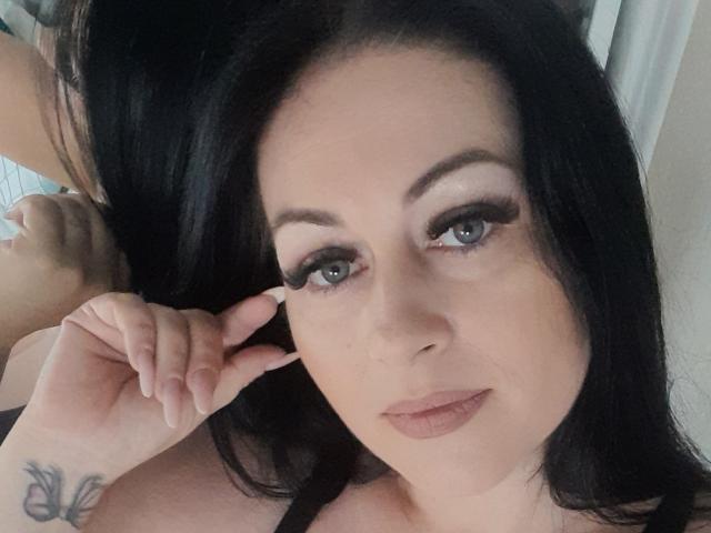 Sellhena mit schwarzen Haaren, blauen Augen und langen Wimpern, die ihre Wange mit ihrer rechten Hand berührt und dabei ein Schmetterlingstattoo an ihrem Handgelenk zeigt. Ihre Brüste sind teilweise sichtbar..