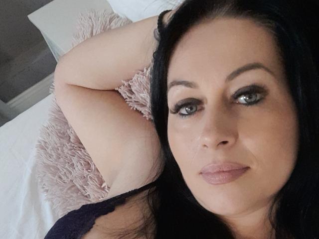 Sellhena, con capelli neri, occhi verdi e rossetto rosa, sdraiata su un letto con un cuscino rosa soffice, braccia dietro la testa, indossa una maglia scura in pizzo e guarda verso la fotocamera..