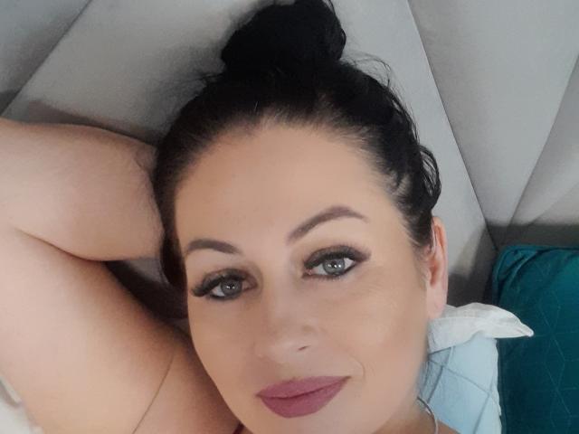 El rostro de Sellhena, acostada, con cabello oscuro recogido en un moño, ojos verdes, cejas gruesas, pestañas largas, lápiz labial rosado-rojizo, brazo detrás de la cabeza, piel clara, ligera sonrisa, sobre ropa de cama blanca y turquesa..