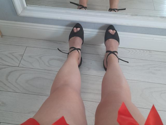 Las piernas de Sellhena, con tacones negros de punta abierta y detalles de corazones recortados, están sobre un suelo de madera clara, y se reflejan en un espejo. Su falda roja es visible en parte en la parte inferior..