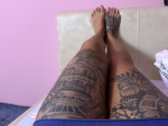 AndraD's tattooed thighs with detailed black ink, showing a building and butterfly on left and right respectively, crossed at the ankles, pink toenails, lying on a bed.