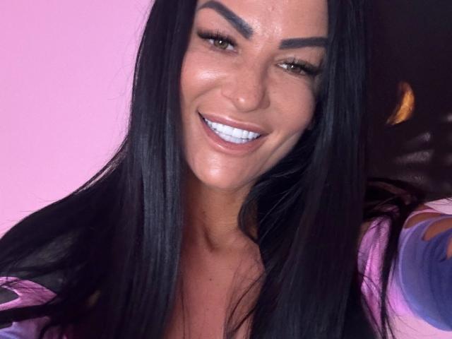 AndraD has shiny black hair, thick eyebrows, and a bright white smile with visible teeth. She's wearing a pink and purple top, and her skin is tan.