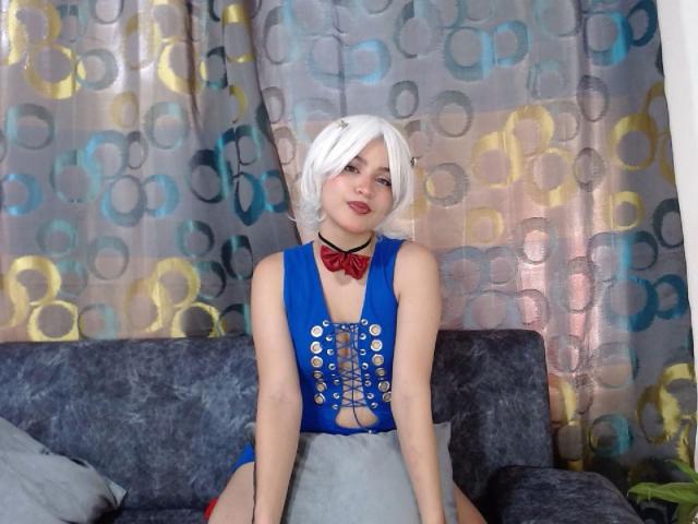CollinsCharlotte with white bob, wearing blue cutout dress, red bow tie, sitting on gray couch, showing her tits through dress, smiling, white skin, red lipstick, pale makeup.