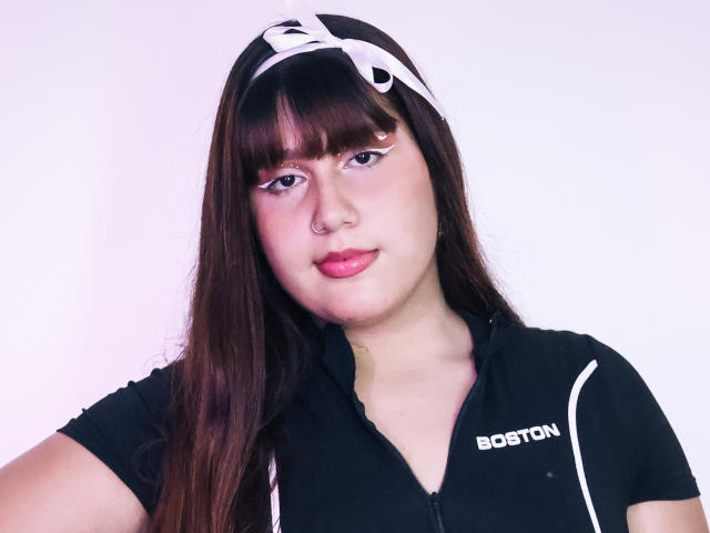 BibiCruz with long brown hair, white eyeliner, pink lipstick, black zip-up shirt, white headband, nose ring, and "Borton" logo, looking at camera with a slight smile.