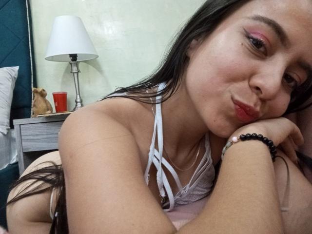 JessyLenon with pink eyeshadow, white strappy top, pouty red lips, black hair, resting on bed, right arm bracelet, left arm on pillow, red cup in background.