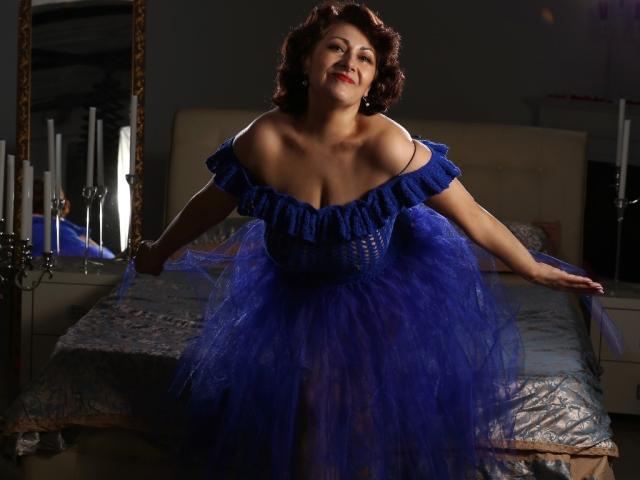 SophiaLaur in a blue off-shoulder dress with a fluffy purple tutu, leaning forward, smiling, showing her big tits, with curly brown hair, in a dimly lit room.