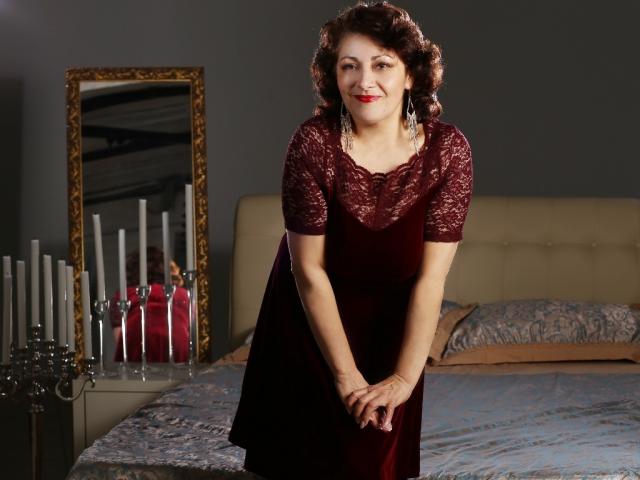 SophiaLaur, uma mulher curvilínea com batom vermelho e cabelos castanhos cacheados, vestindo um vestido de renda escuro, inclinada para frente com as mãos juntas, em pé ao lado de uma cama com velas e um espelho..