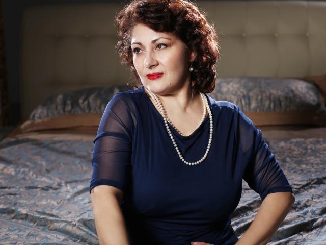 SophiaLaur, with curly brown hair and red lipstick, wears a sheer blue top, pearl necklace, and looks slightly to the side. Her tits are modest, her ass is unseen.