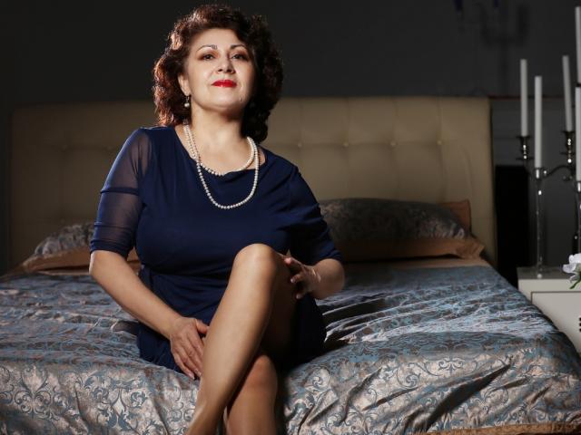 SophiaLaur, una mujer curvilínea con senos de tamaño medio, vestida con un vestido azul marino y un collar de perlas, sentada en una cama con sábanas estampadas, con las piernas cruzadas, lápiz labial rojo y cabello castaño rizado..