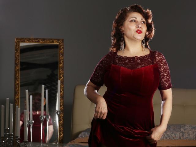 SophiaLaur, con i capelli castano ricci, rossetto rosso, indossa un abito rosso in pizzo, in piedi, tiene l'abito, riflesso in uno specchio decorato, candele bianche sul letto, parete grigia..