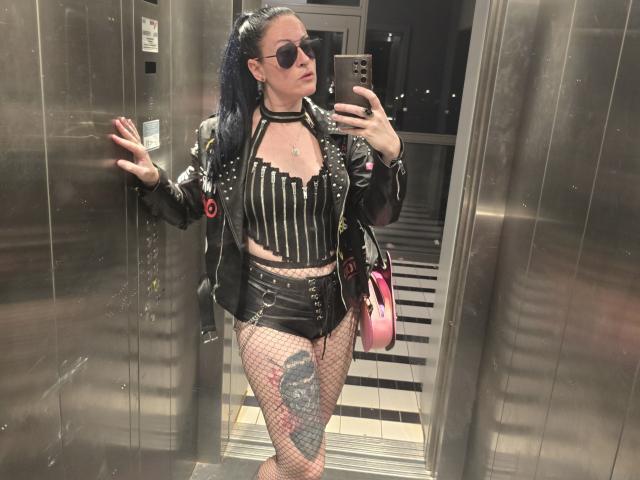 GennyRock, con due codini neri, indossa una giacca di pelle con borchie, una maglietta a righe, pantaloncini neri, collant a rete e una collana. Sta scattando un selfie allo specchio in un ascensore. Il suo tatuaggio è visibile..