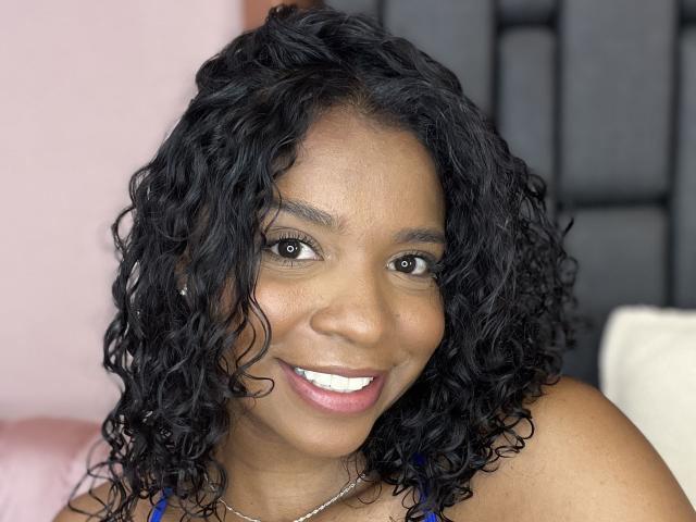 LisaMendez has dark curly black hair, brown skin, and a cute smile with white teeth. She's wearing a blue top and a silver necklace, looking happy and relaxed.