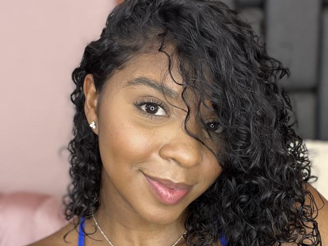 LisaMendez ha i capelli ricci e scuri, gli occhi marroni e un sorriso carino con il rossetto rosa. Indossa una maglietta blu e piccoli orecchini di diamanti. La sua pelle è liscia e marrone..
