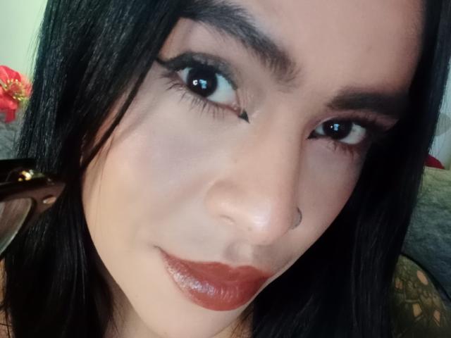 StellaBeckker's face, dark straight hair, brown eyes with thick black eyeliner, nose ring, glossy red lipstick, holding a brown pen on the left side.