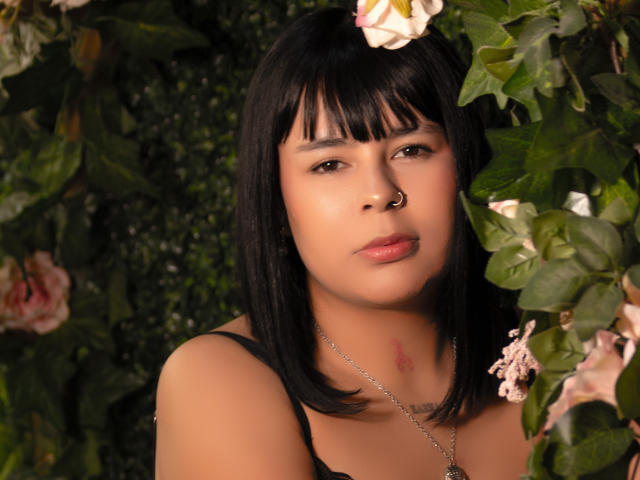 KylieDark with black bob, nose ring, and pink flowers in her hair, wearing a black top, showing a small bruise on her neck, looking at the camera.