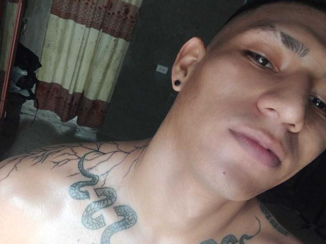 Peter Jonies, ohne Hemd, hat ein Schlangentattoo auf seiner Brust, schwarze Ohrlöcher und leicht geschlossene Lippen. Kurze Haare, gebräunte Haut und braune Augen. Der Hintergrund ist dunkel und unscharf..