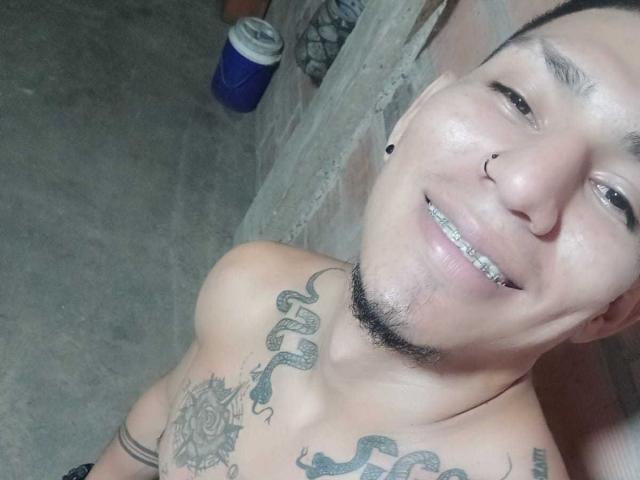 PeterJonies, sin camisa y con tatuajes, sonriendo a la cámara, con un piercing en la nariz, barba de tres días, perforaciones en las orejas y una taza de plástico azul en el fondo. PeterJonies tiene un tatuaje visible del pene..